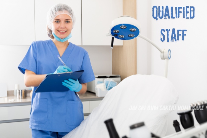 A smiling healthcare professional in blue scrubs and gloves, holding a clipboard in a well-equipped medical environment. This image showcases the qualified staff at Jai Sri Ohm Sakthi Home Care in Coimbatore, highlighting their commitment to providing expert care and support in home nursing services for patients.